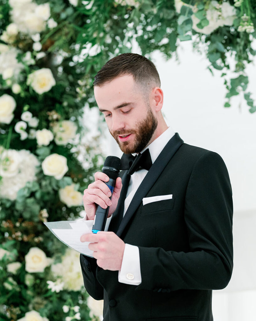 Man giving a speech at a wedding ceremony with a floral arch in the background.