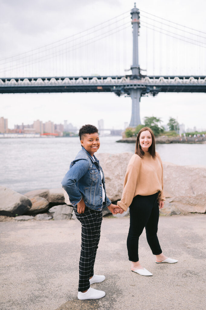 Couple holding hands and smiling while walking against a view of the Manhattan Bridge. 