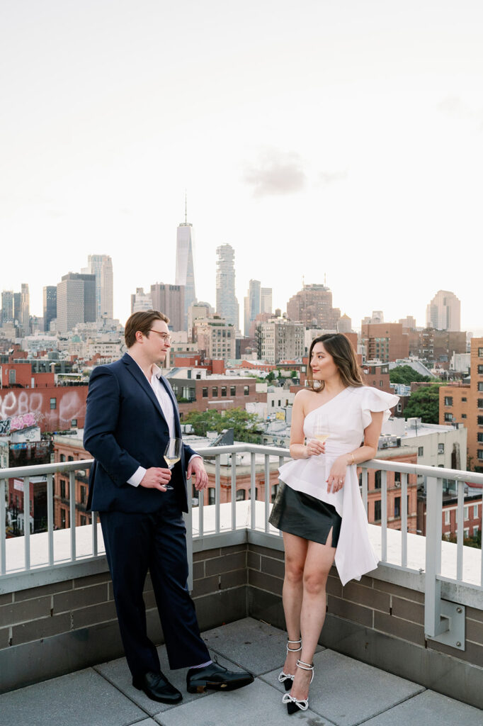 Chic couple casually drinking a glass of wine on a rooftop in Manhattan with views of the city for their engagement photos. 