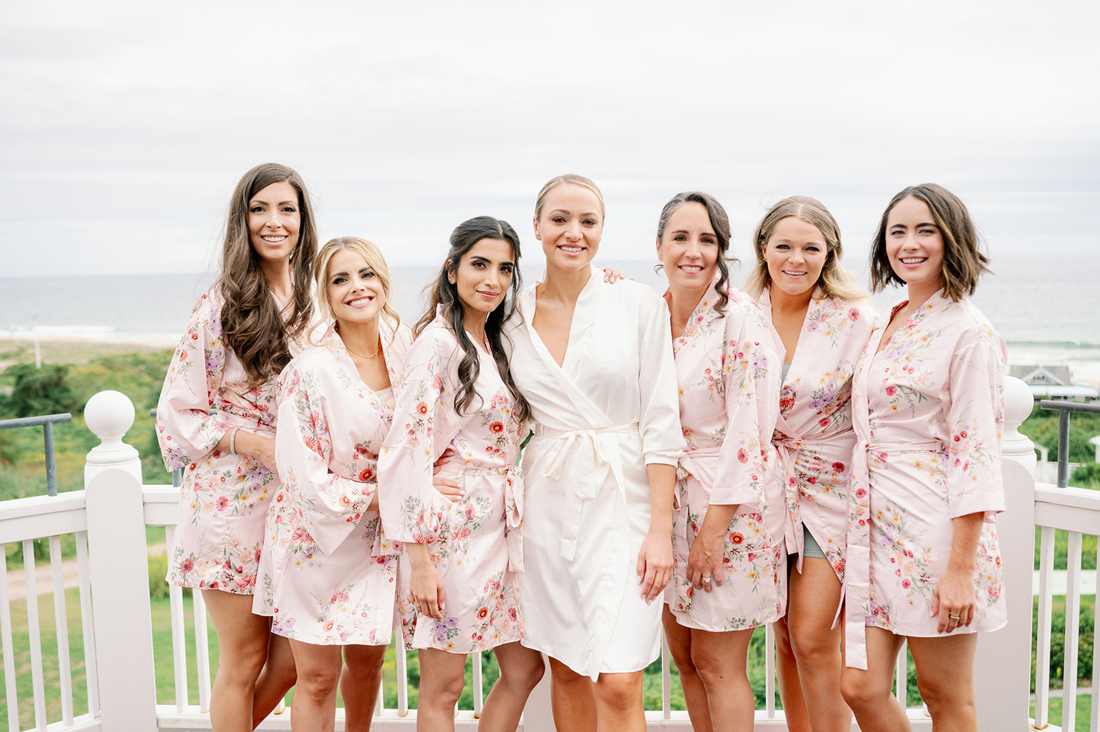 A bride and bridesmaids wearing matching pink floral robes standing on a balcony after getting their hair and makeup done. 