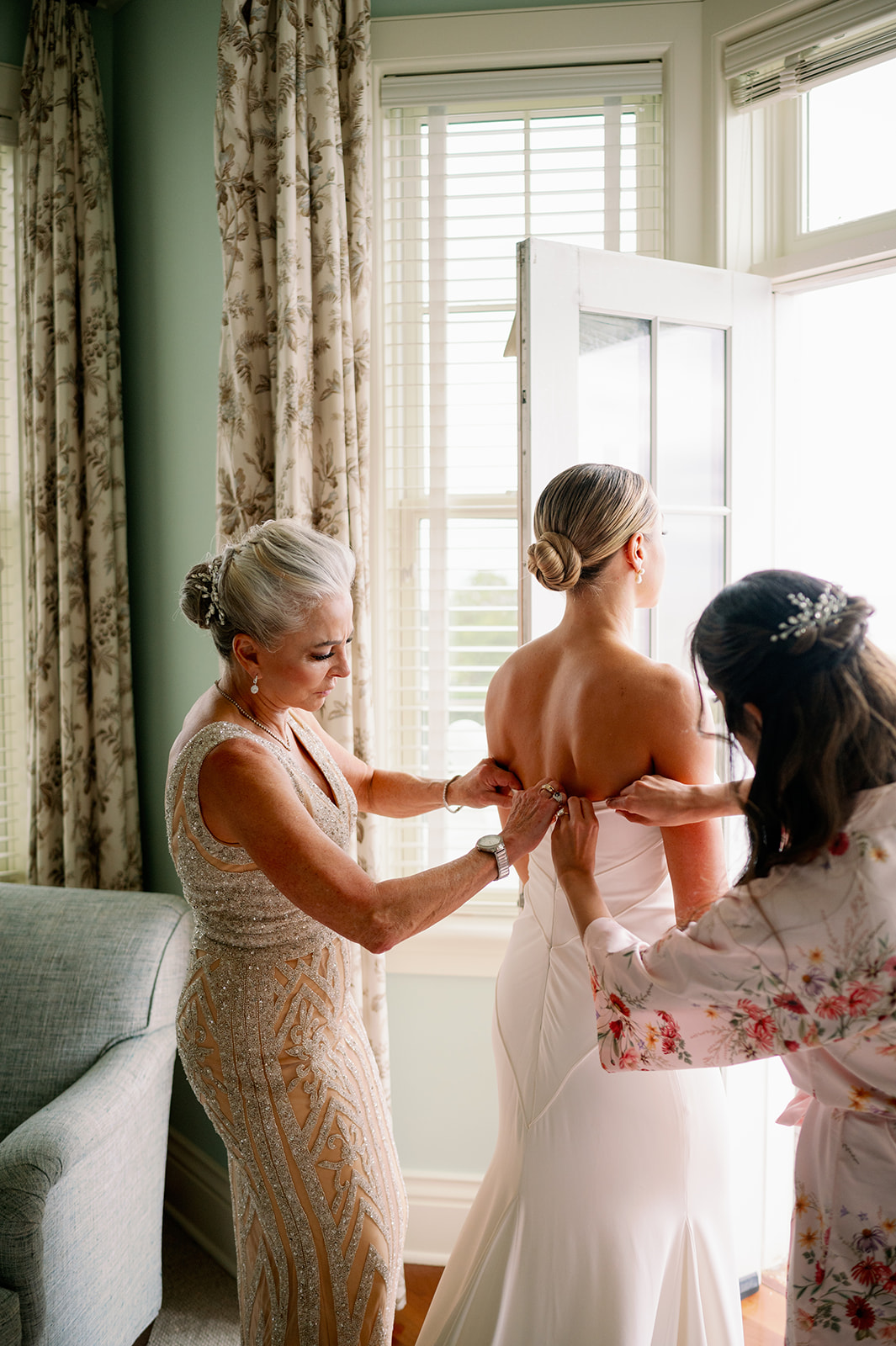 A New England bride getting her wedding dress zipped up by her mother and mother-in-law. 