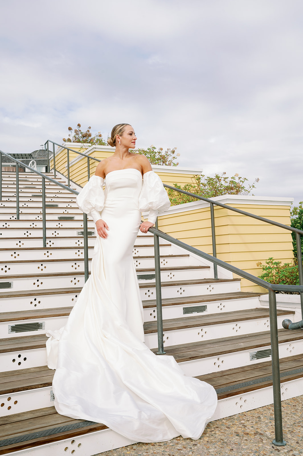 A stunning modern bride posing on the steps of Ocean House wearing a gorgeous strapless dress with a puff sleeve cape. 
