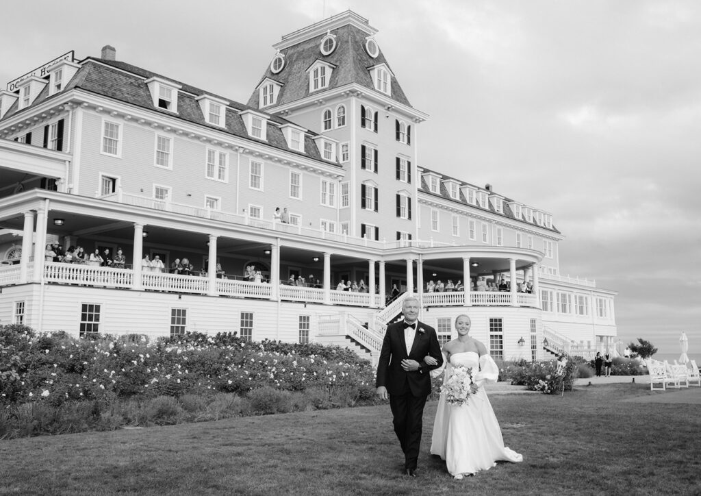 Black and white capture of a bride and her father walking down the aisle with Ocean House in the Background 