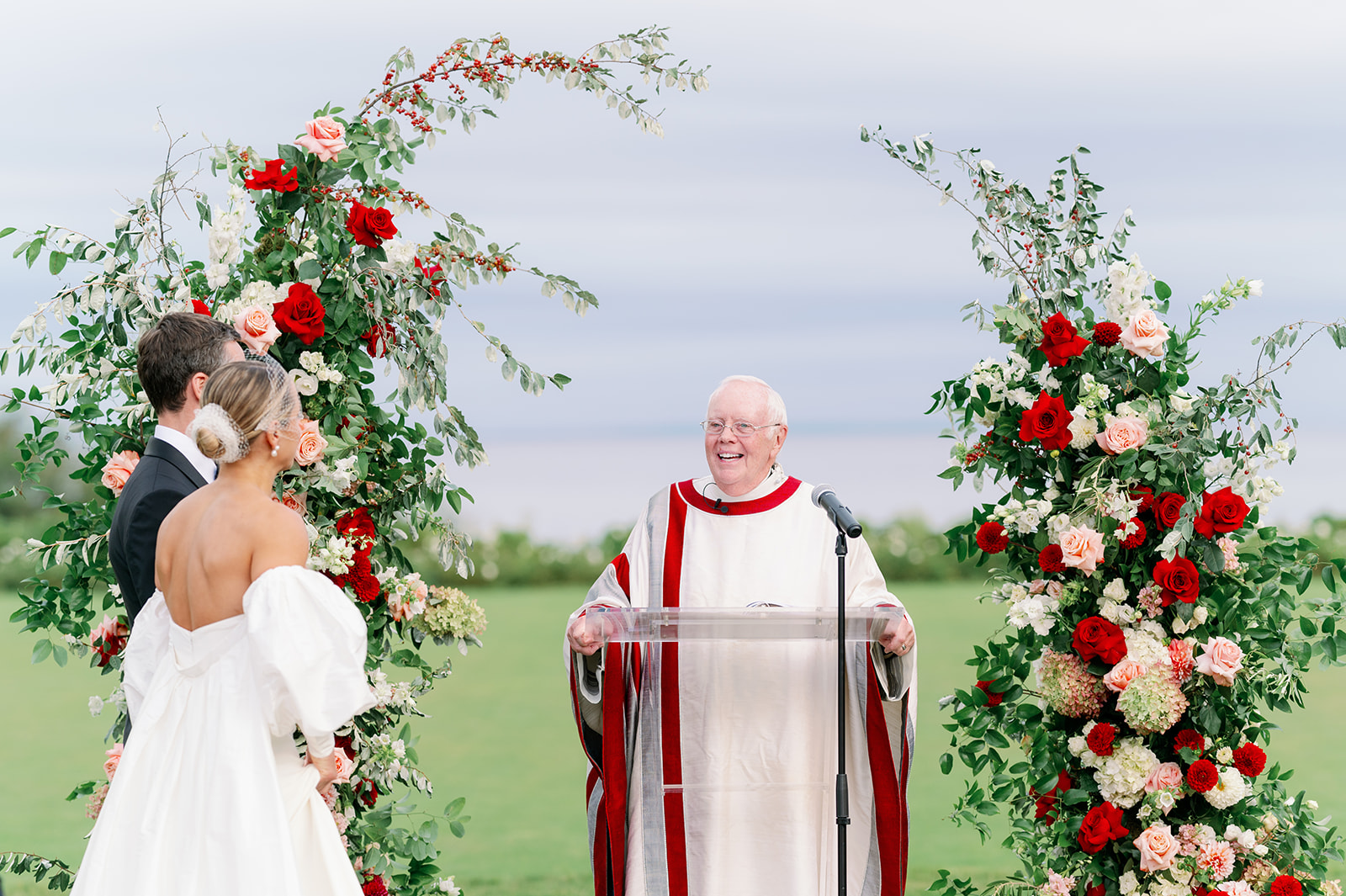 A bride and groom at the altar of their outdoor seaside wedding ceremony in Rhode Island with a floral arch and catholic priest. 