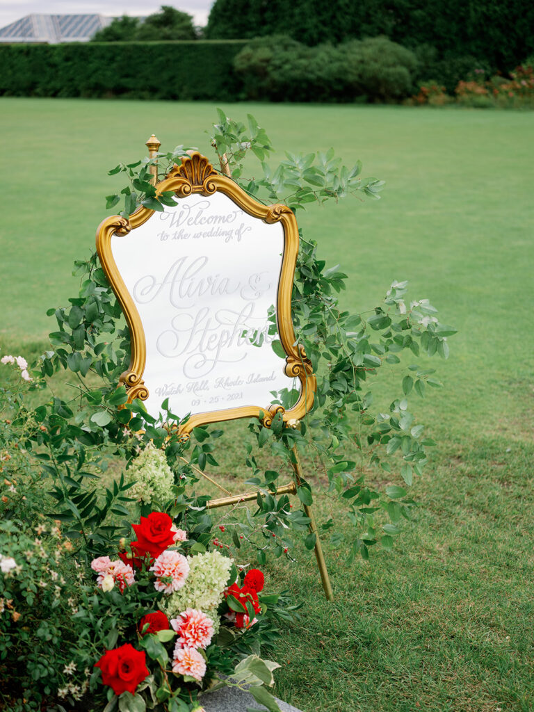 Elegant gold mirror wedding welcome sign on an easel surrounded by greenery and a red and pink floral arrangement. 