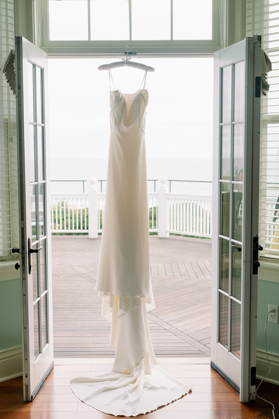 A stunning wedding gown with a long train hanging from a balcony door frame at Ocean House in Rhode Island with the ocean in the background.