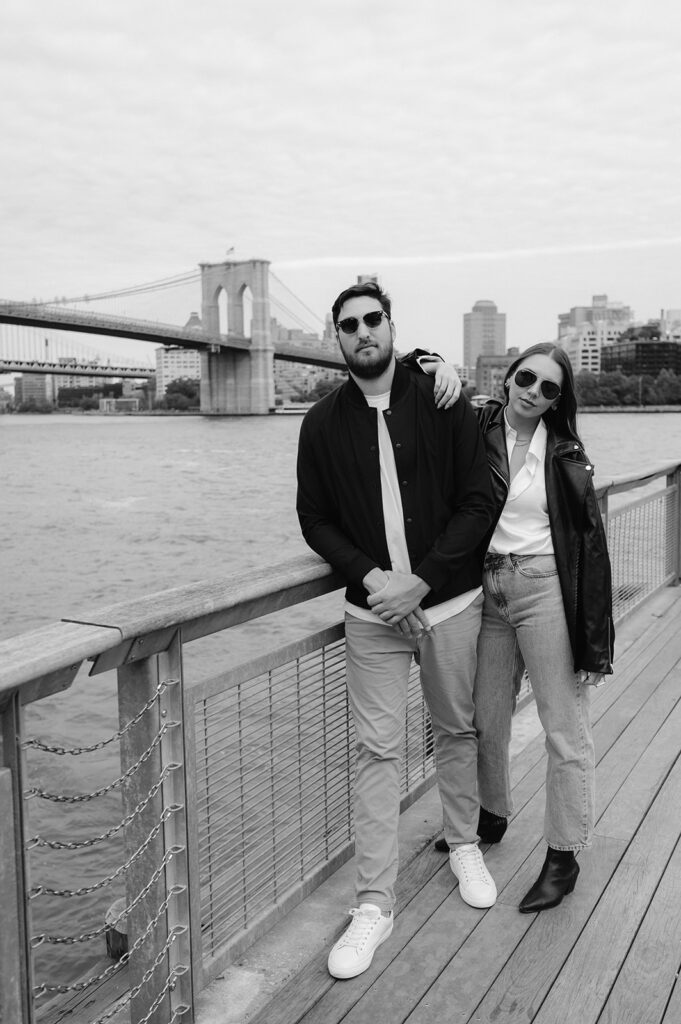Cool couple wearing sunglasses posing for their engagement photos with the Brooklyn Bridge in the background. 