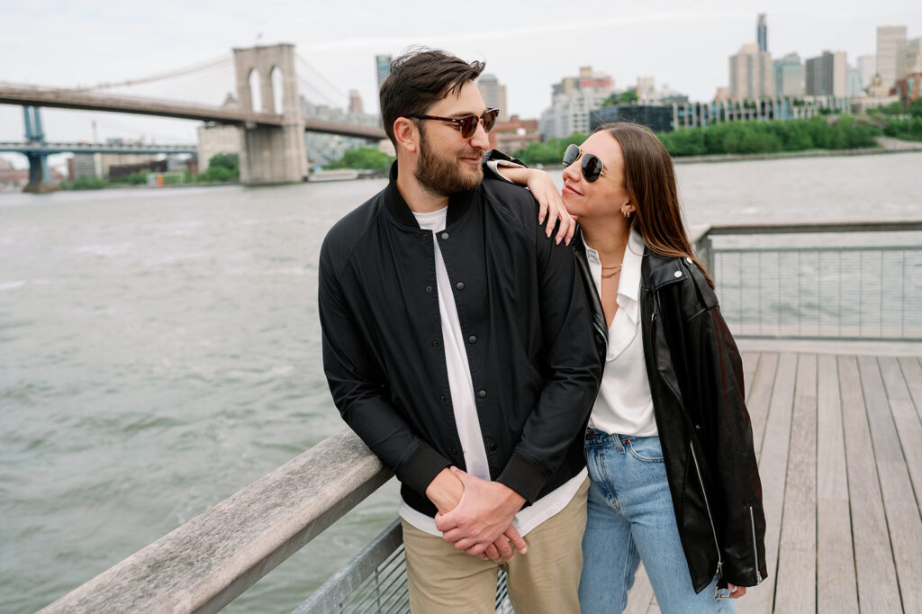 Stylish couple wearing sunglasses leaning on a railing in South Street Seaport overlooking the East River and Brooklyn Bridge. 
