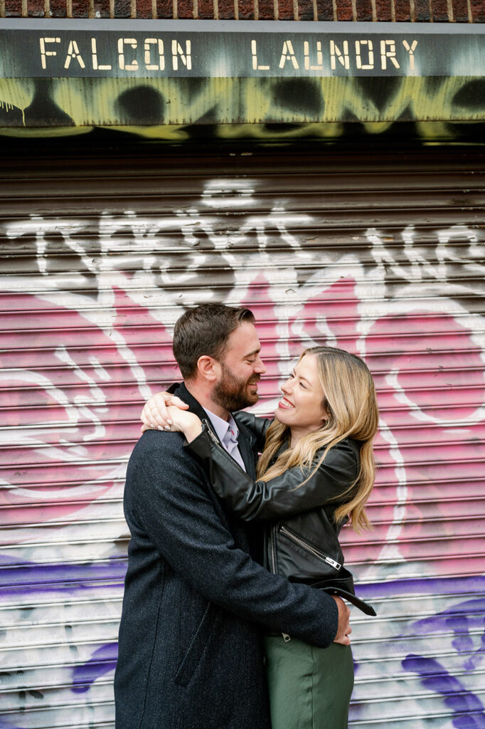 Couple embracing candidly for their urban engagement photoshoot against a graffiti backdrop in Williamsburg, NY. 