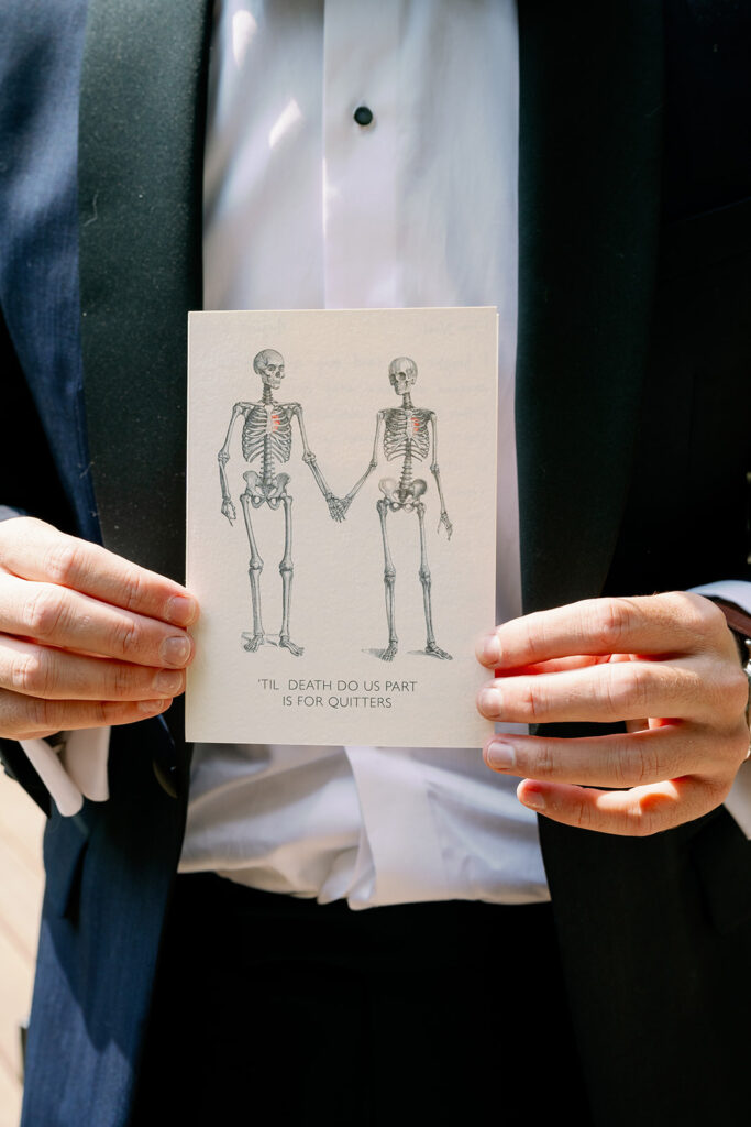 Groom holding up a card with two skeletons holding hands that says "Til death do us part is for quitters."