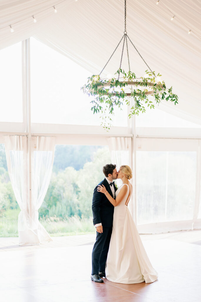 Bride and groom kissing on the dance floor of their tented wedding reception with a greenery chandelier.