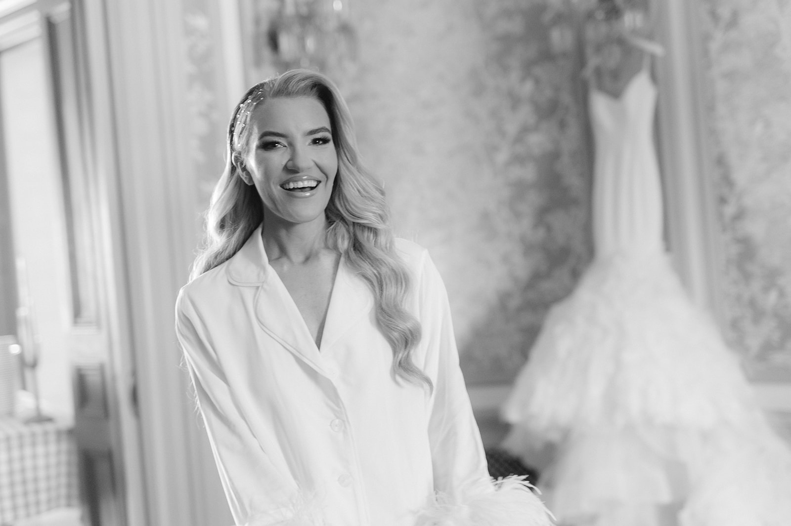 Bride in pajamas smiling with her wedding dress hanging in the background.