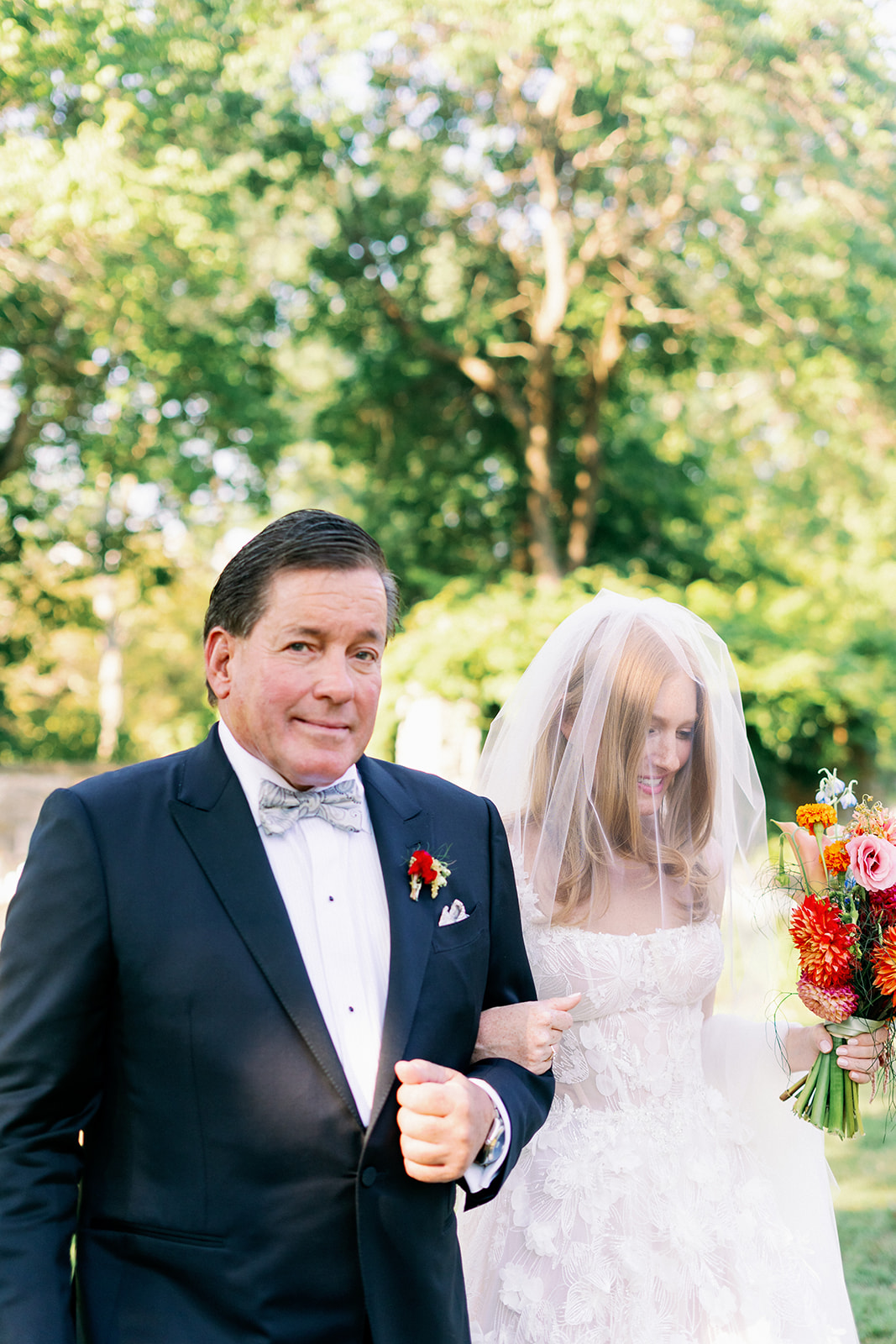Bride walking down the aisle linking arms with her dad and holding a vibrant wildflower bouquet.