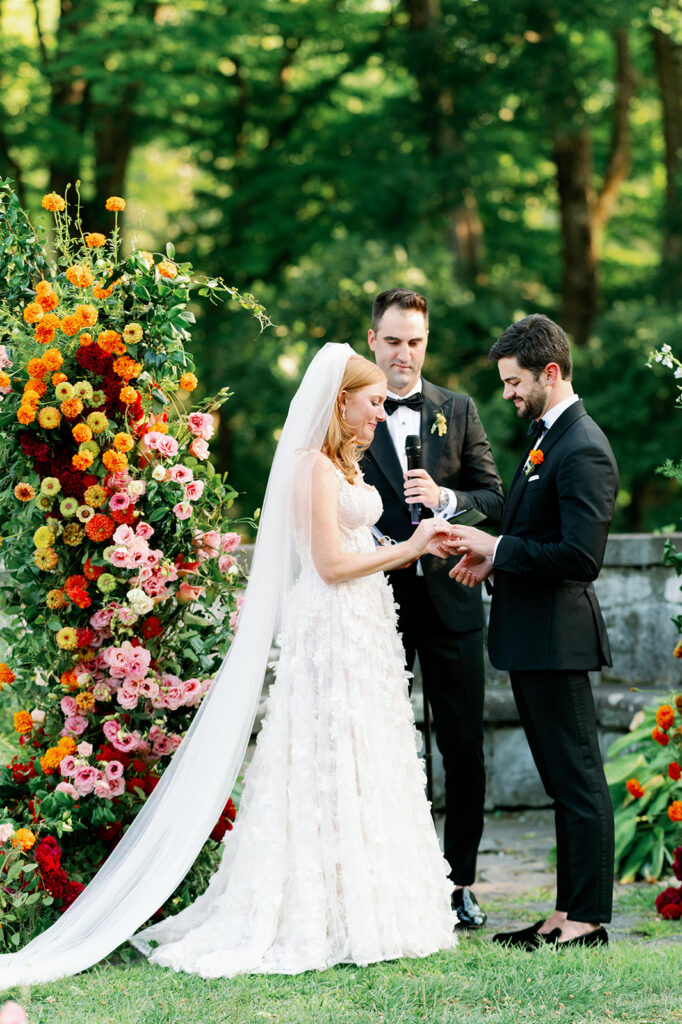 Bride putting on the groom's ring during the ceremony against a vibrant floral backdrop. 