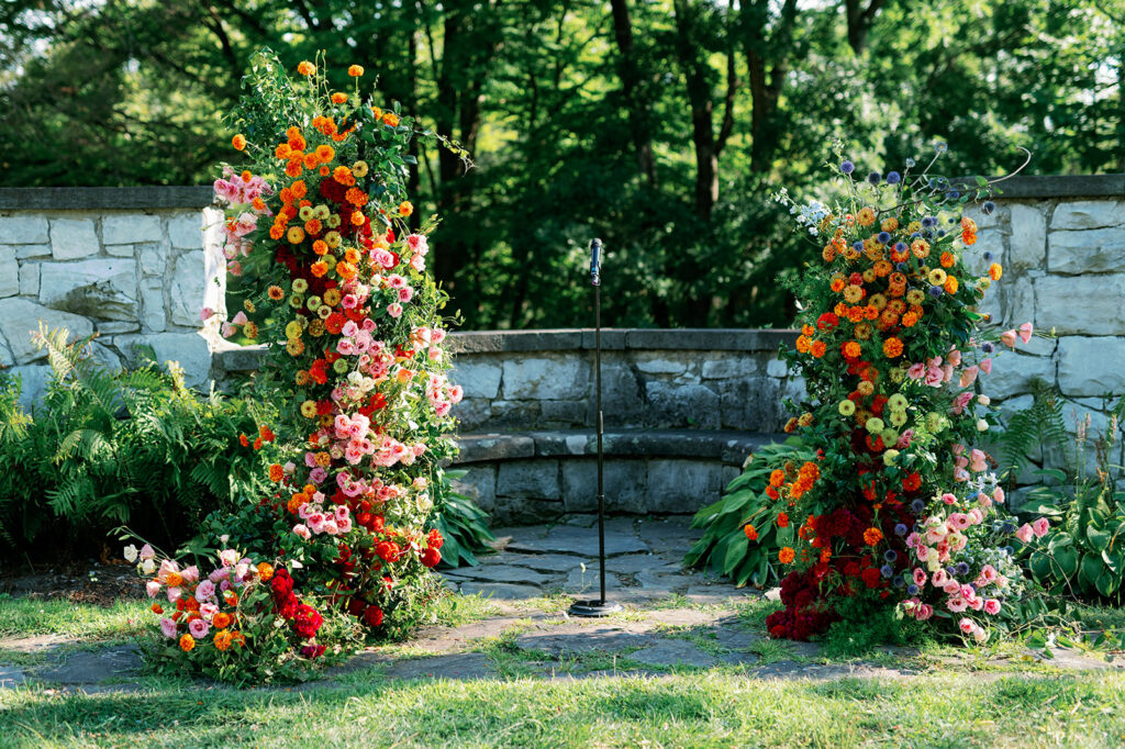 Gorgeous summer wedding ceremony freestanding floral arch backdrop with vibrant orange, red, and pink wildflowers and greenery.