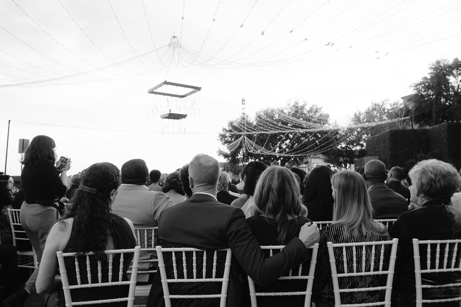 Black and white portrait of wedding guests watching the ceremony under a canopy of fairy lights.