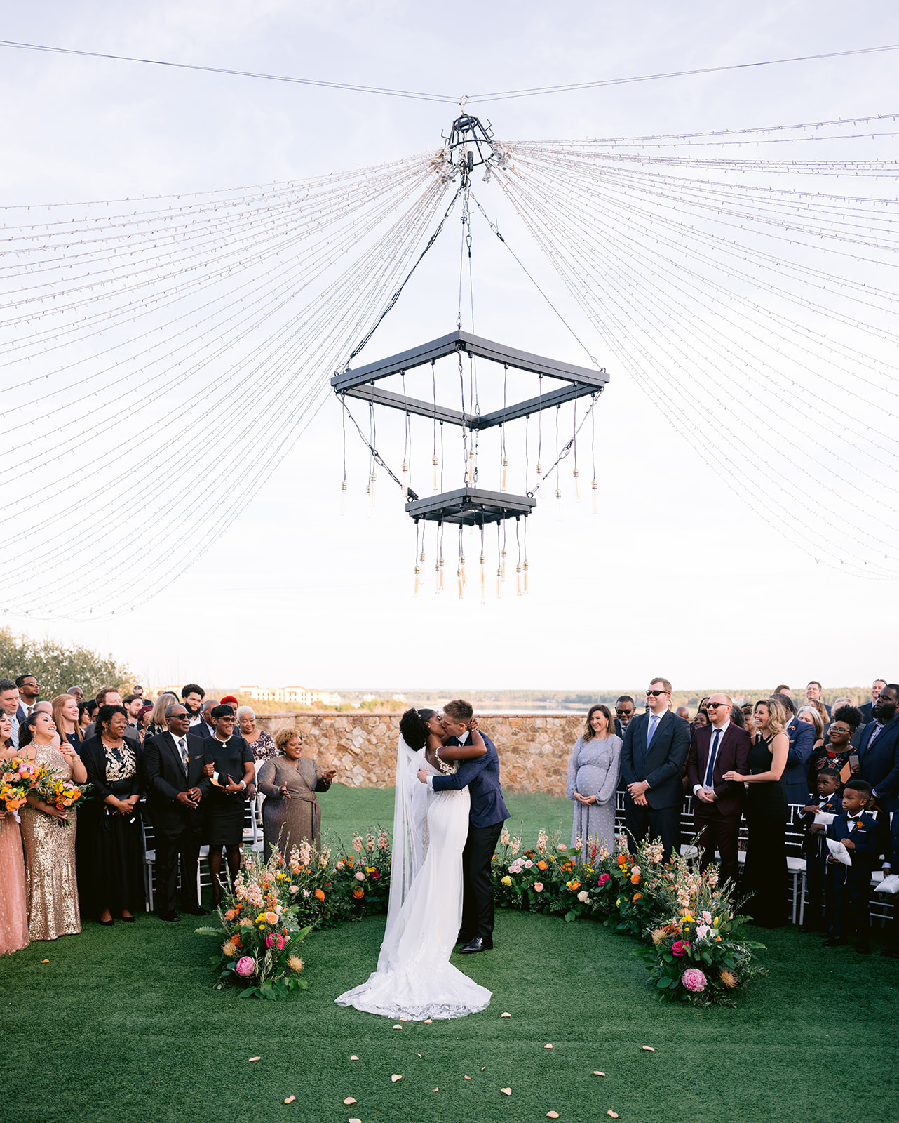 Bride and groom first kiss under a canopy of fairy lights surrounded by vibrant florals.