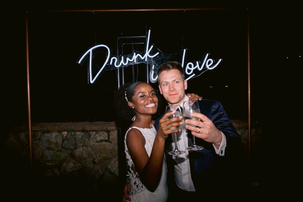Bride and groom tasting under a neon 'drunk in love' sign.