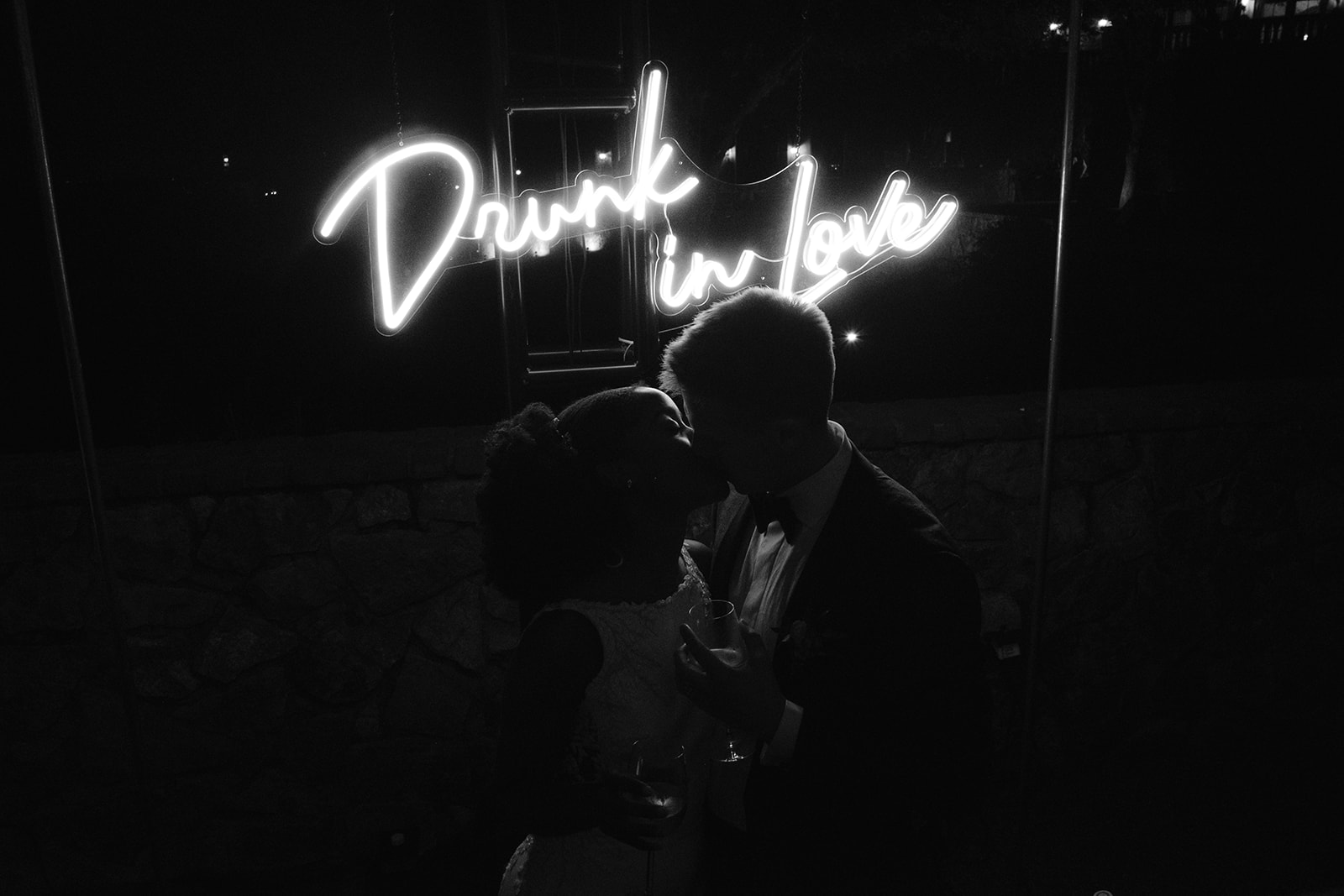 Bride and groom kissing under a neon 'drunk in love' sign.