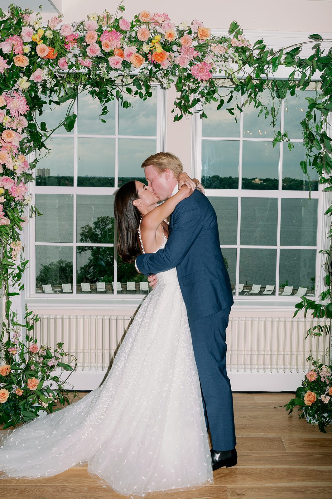 Bride and groom first kiss under a floral arch during their indoor wedding ceremony at Minikahda Club.
