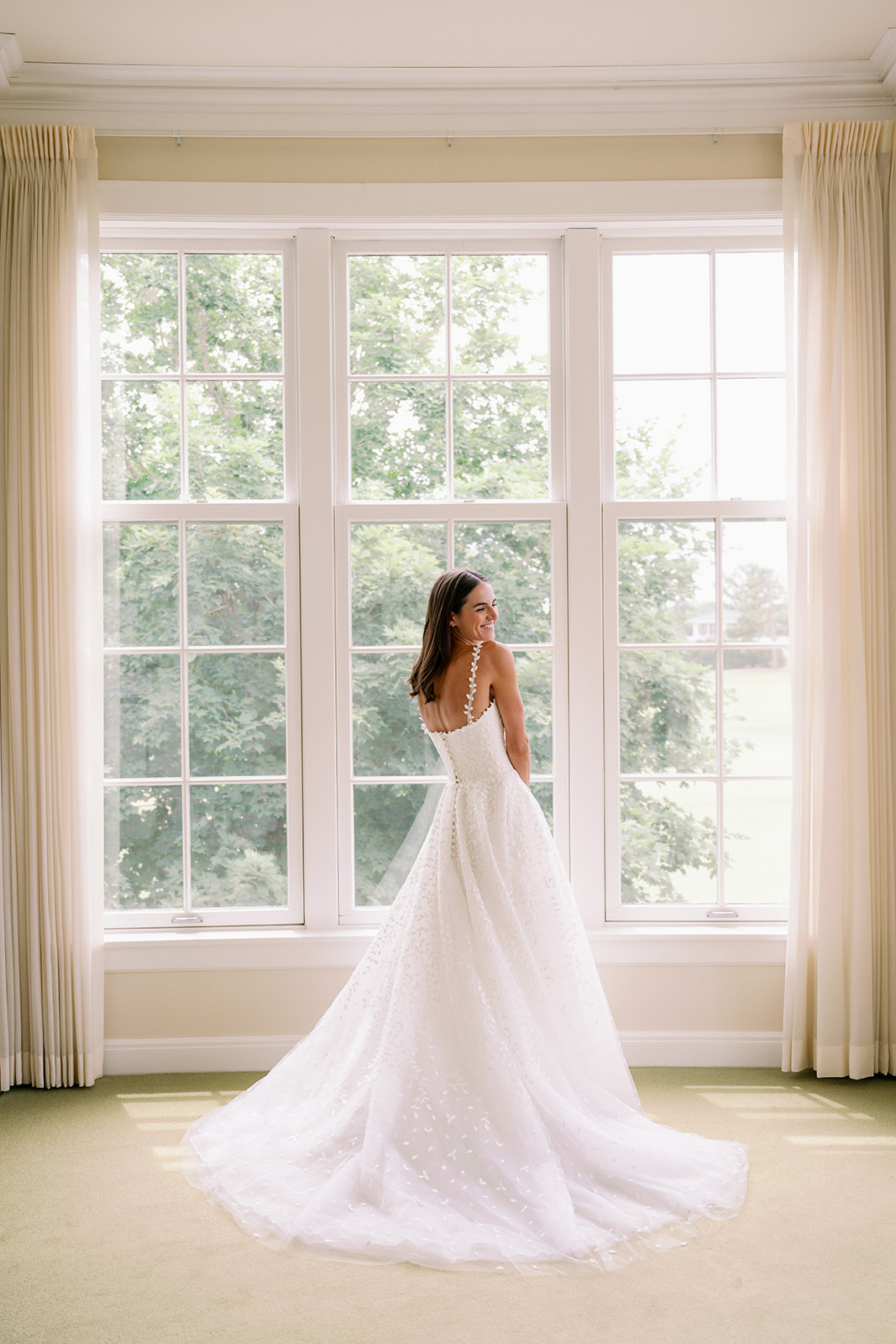 Portrait of Carly in her wedding dress standing in front of a large paned window inside the Minikahda Club bridal suite.