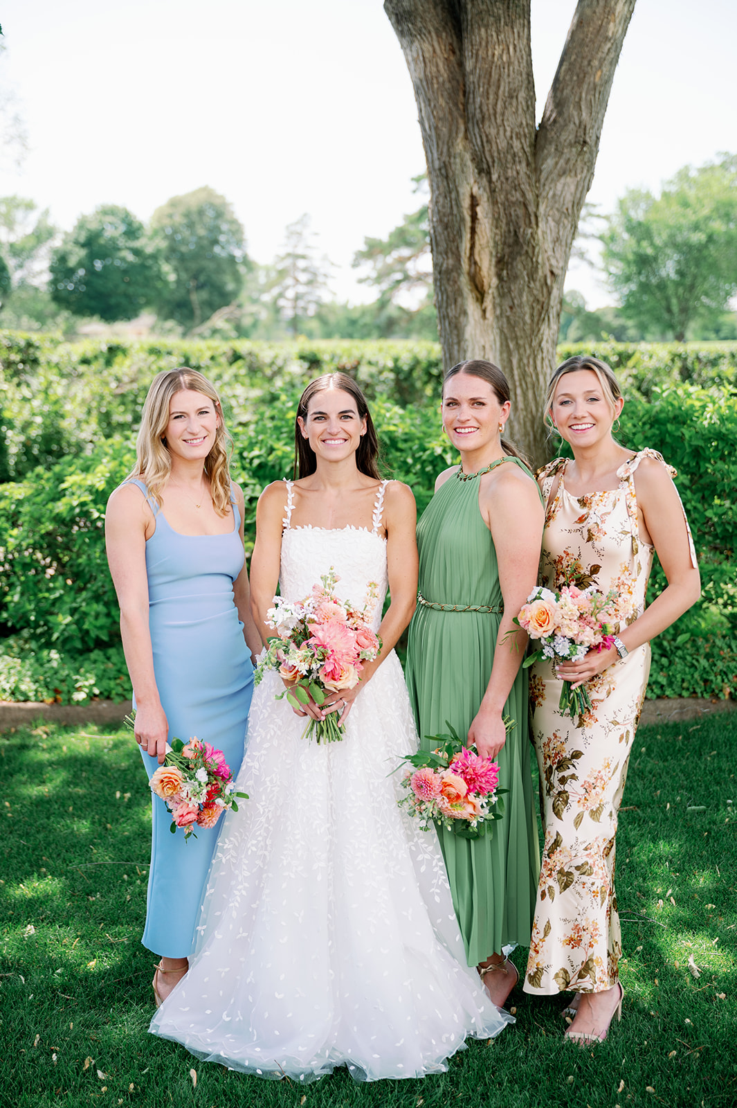Carly and her three bridesmaids in mismatched summer dresses holding pink floral bouquets with a garden backdrop.