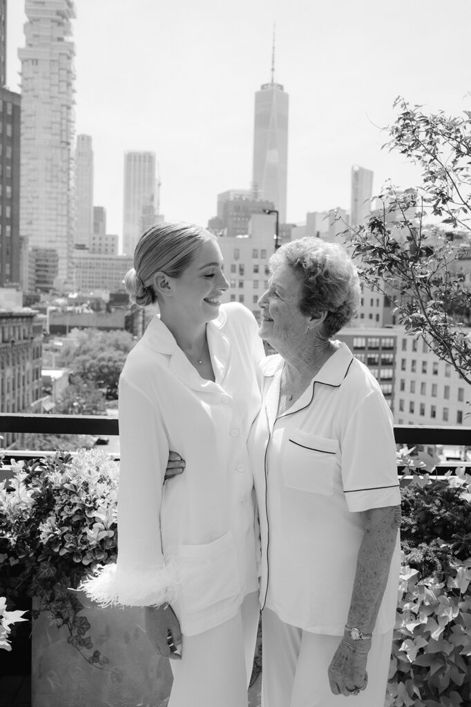 Bride and grandma share smiles in pajamas, cityscape as a backdrop, on the morning of a New York wedding day.