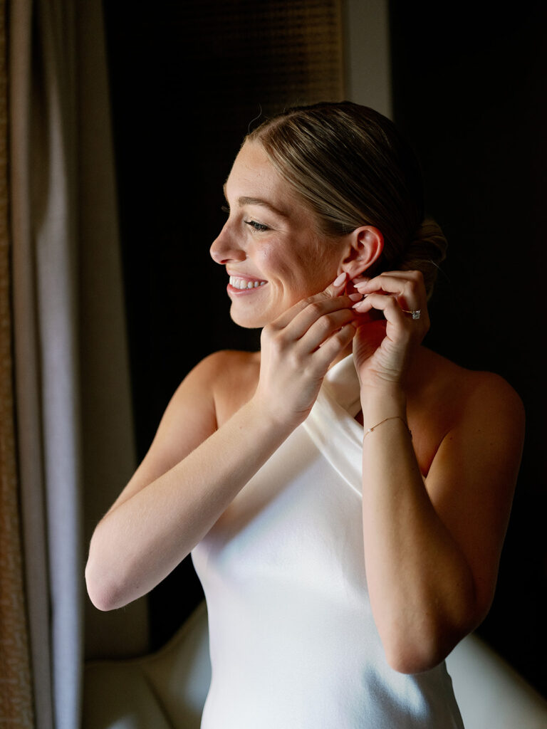 Bride smiles, bathed in natural light, putting on earrings, capturing a moment of grace in a New York wedding morning.
