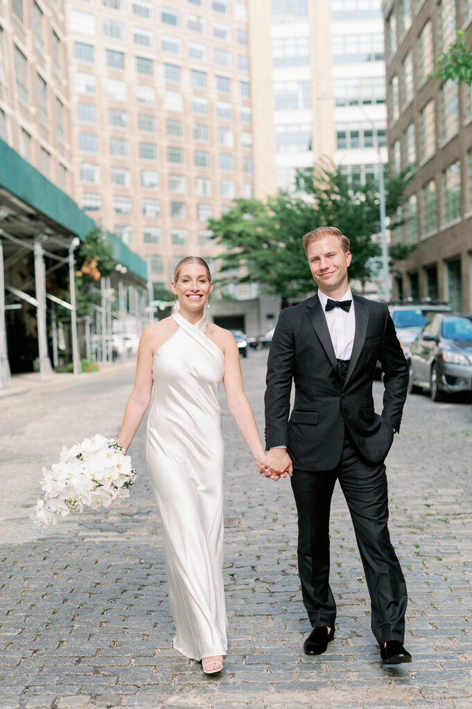 West Village bride and groom walk on a cobblestone street, tall buildings in the backdrop, a candid New York wedding moment.
