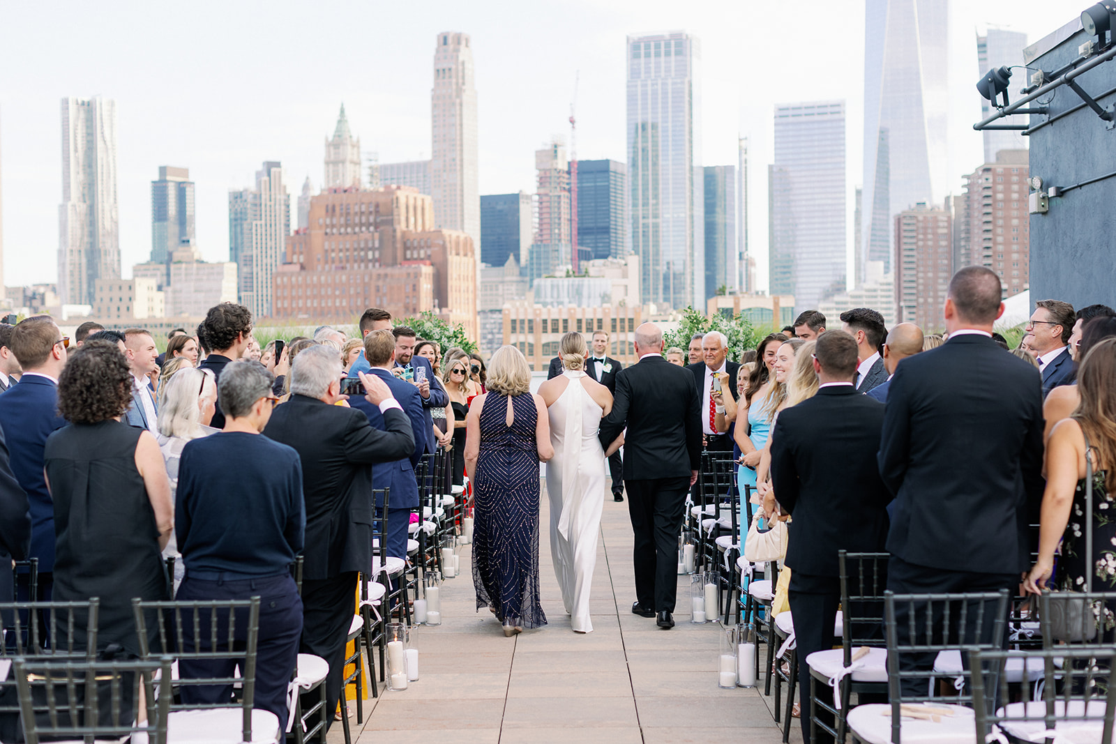 Tribeca Rooftop wedding showcases a wide shot of the bride, joined by her parents, walking down the aisle with stunning city views.