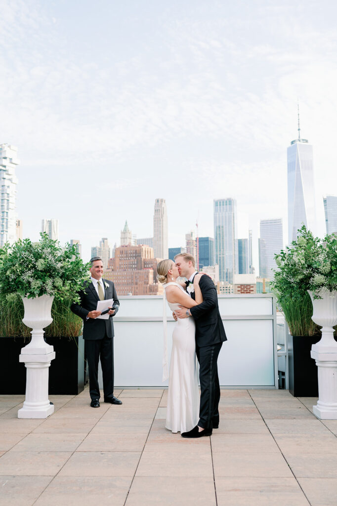 Tribeca Rooftop wedding seals vows with the bride and groom's first kiss, officiant to the side, framed by stunning New York views.