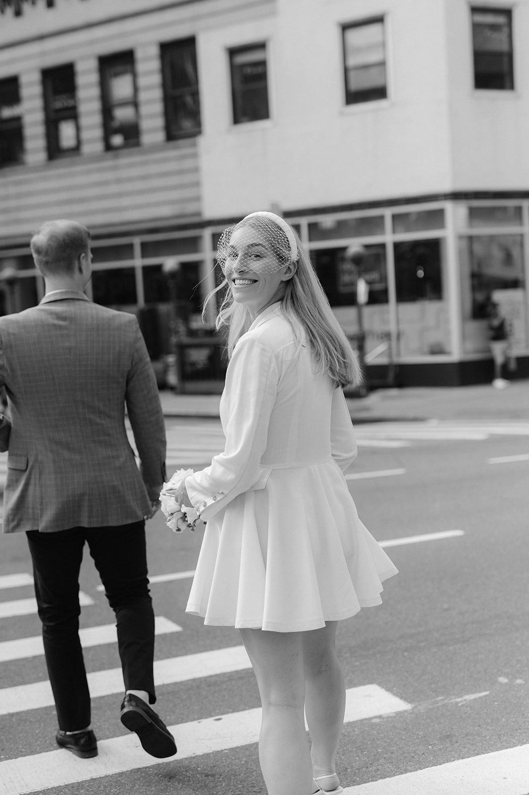 Bride and groom share a candid moment crossing a New York crosswalk, the bride's joyful smile adding charm to this urban love story.