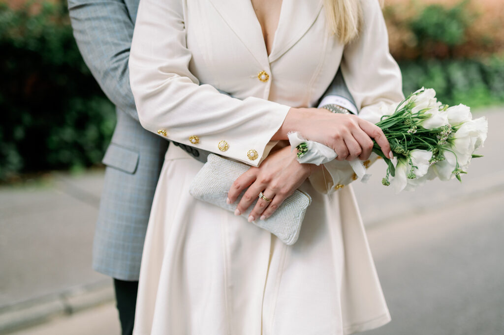Bride, in a vintage suit dress, holds a white bouquet and clutch, embraced by the groom in this intimate New York wedding detail shot.