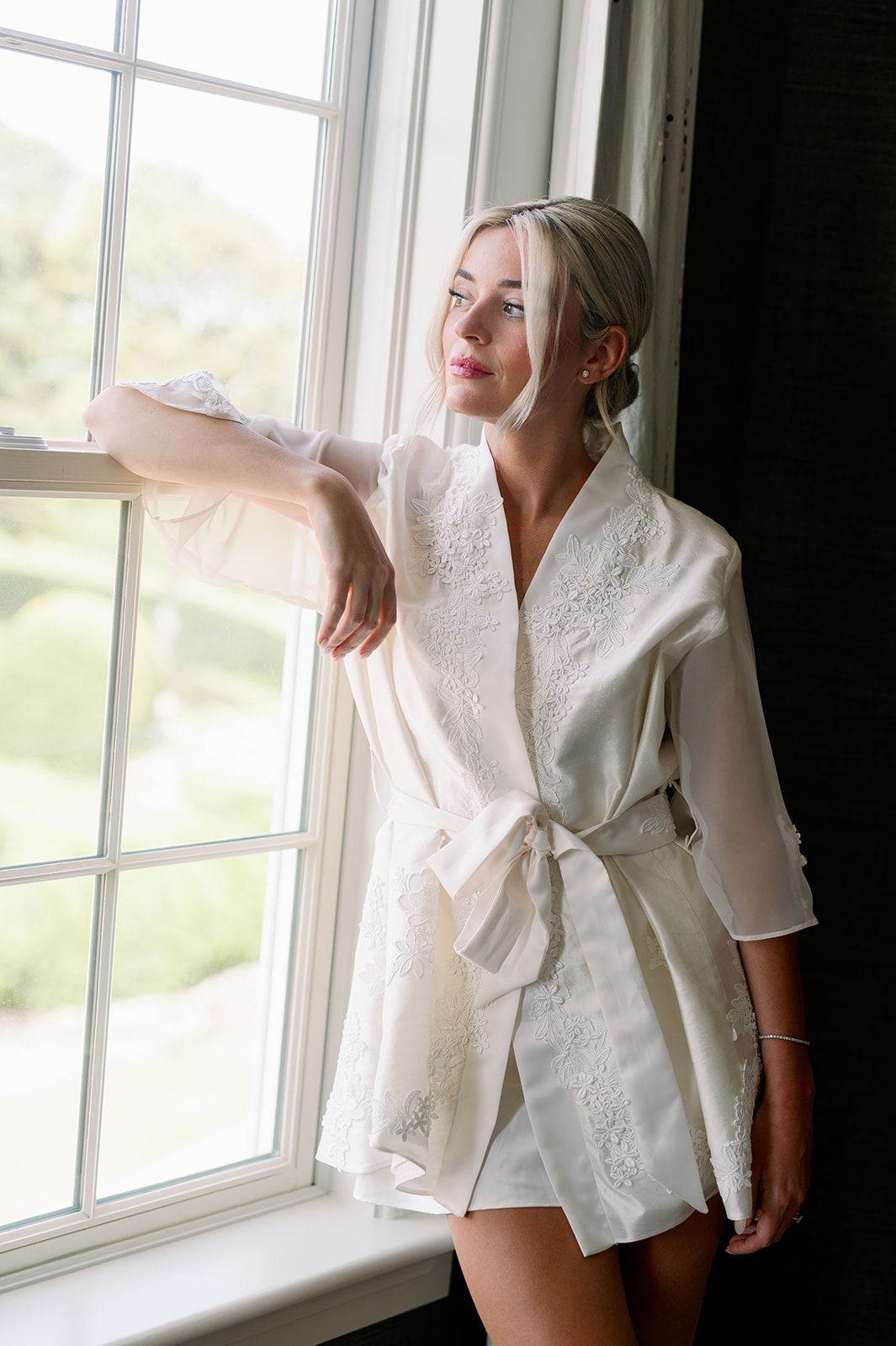 Bride in a custom white robe made from her mom's vintage wedding dress gazing out of a window. 