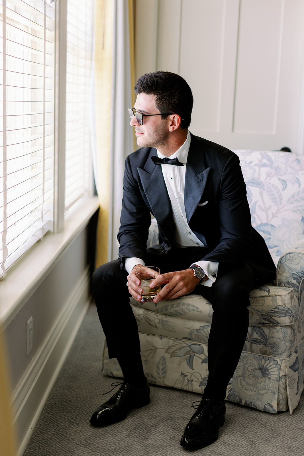 Groom wearing sunglasses and sitting on a chair gazing out of his Ocean House hotel room window. 