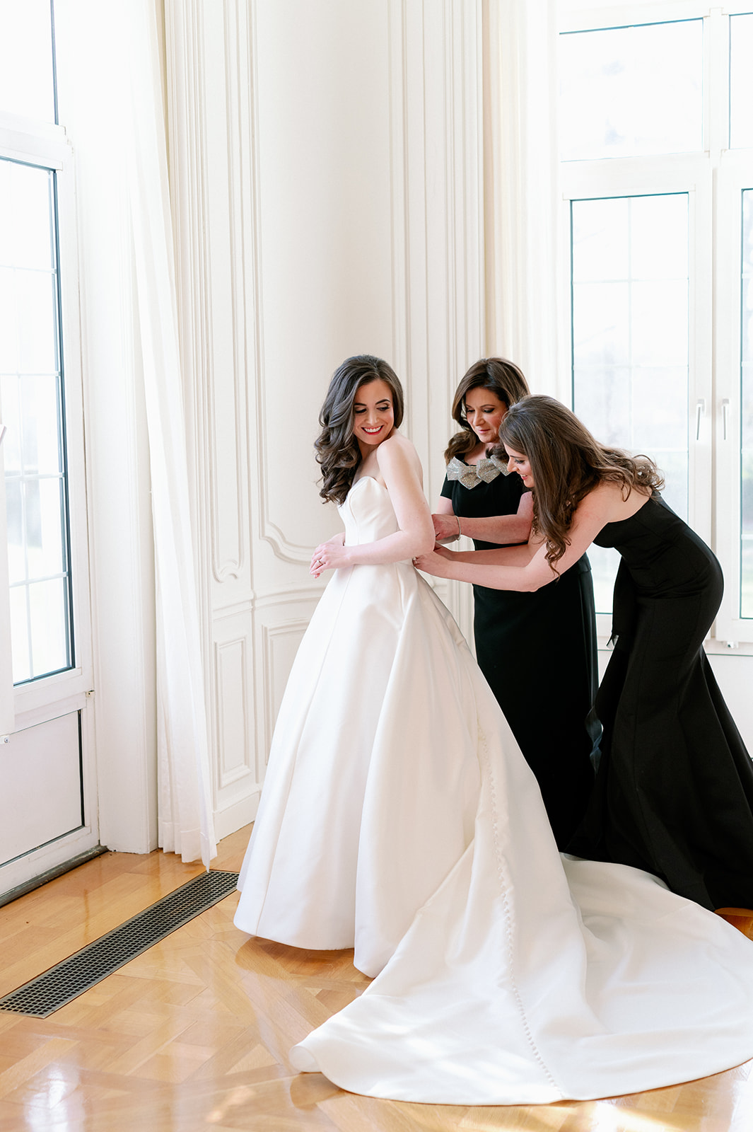 Rachel getting her wedding dress zipped up by her mom and sister in front of large windows at Pine Hollow Country Club.