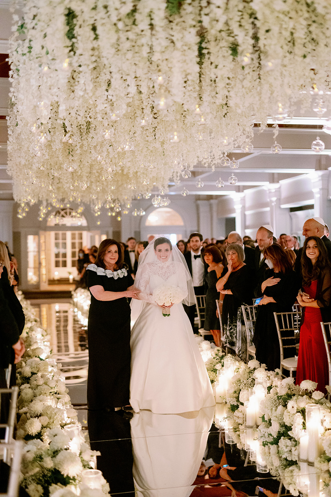 Bride smiling while walking down a reflective, floral-lined aisle with her mom at Pine Hollow Country Club.