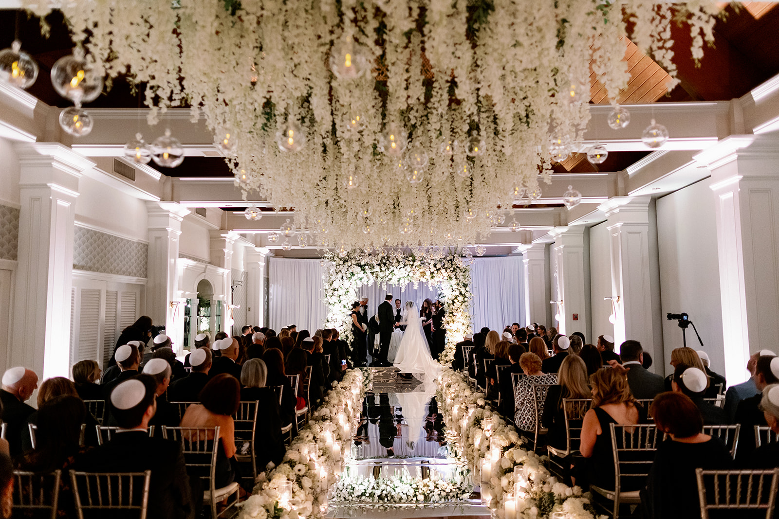 Wide angle view of a luxury indoor wedding with a reflective aisle and hanging florals at Pine Hollow Country Club.