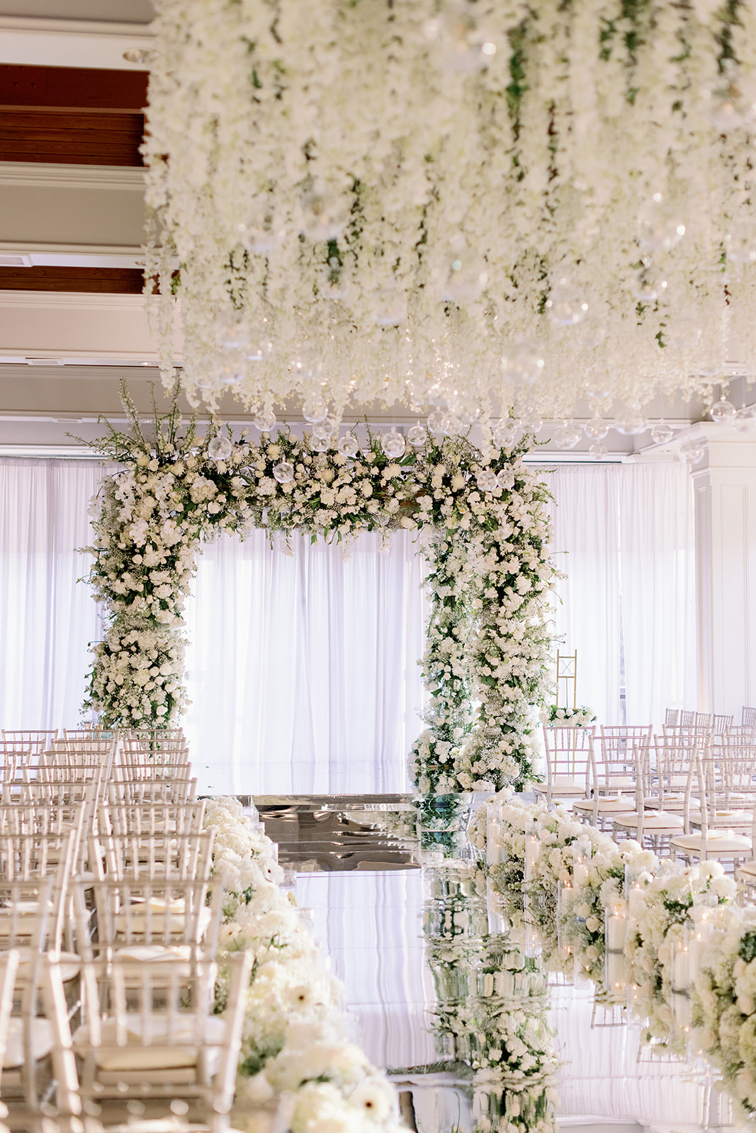 Indoor white floral wedding ceremony with a reflective floor and large floral chuppah.