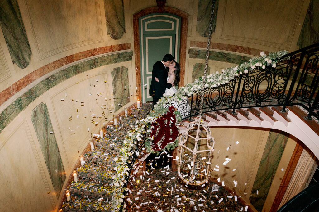 Wide angle view of bride and groom kissing on a spiral staircase with falling confetti. 