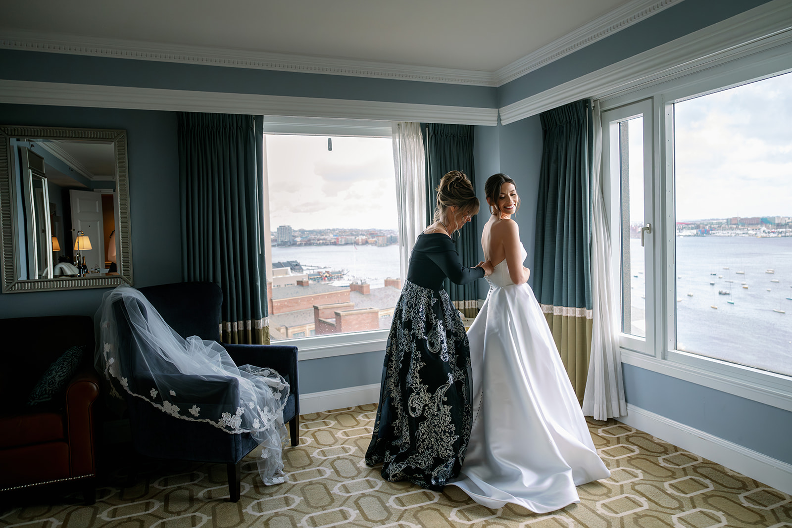 Bride getting her wedding dress buttoned by her mom with panoramic views from the Boston Harbor Hotel. 