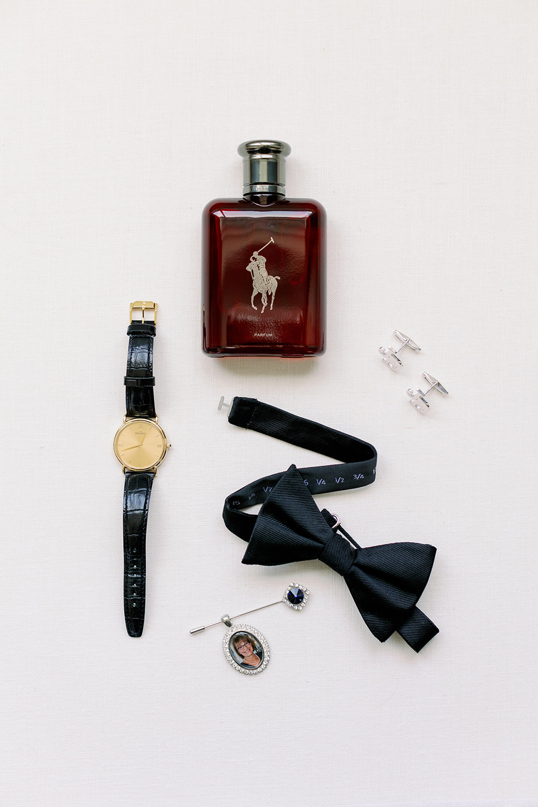 Groom wedding detail flat lay with cologne, watch, bowtie, cufflinks and custom photo pin.