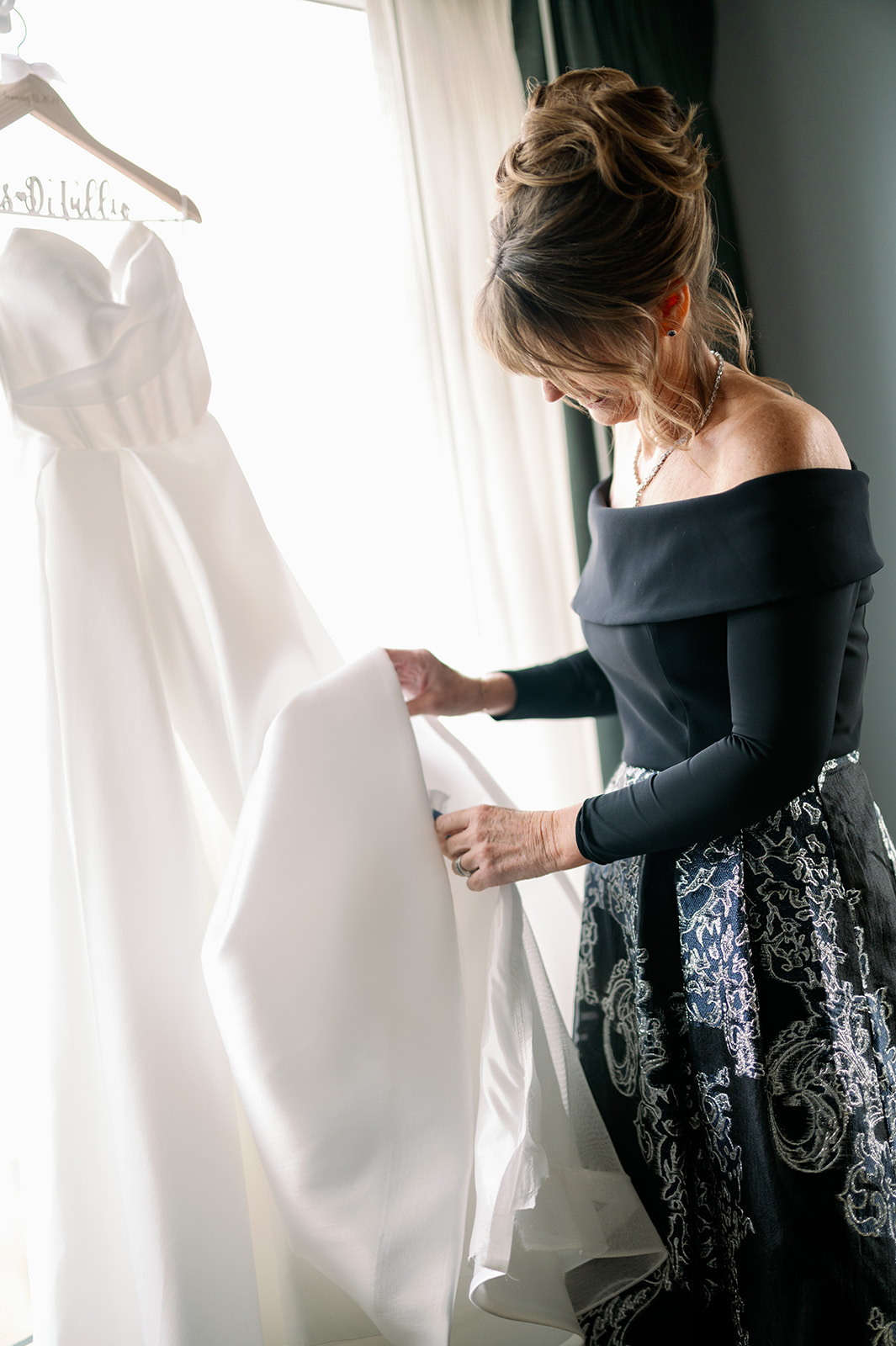 Mother of the bride admiring the wedding dress hanging from brightly lit window. 