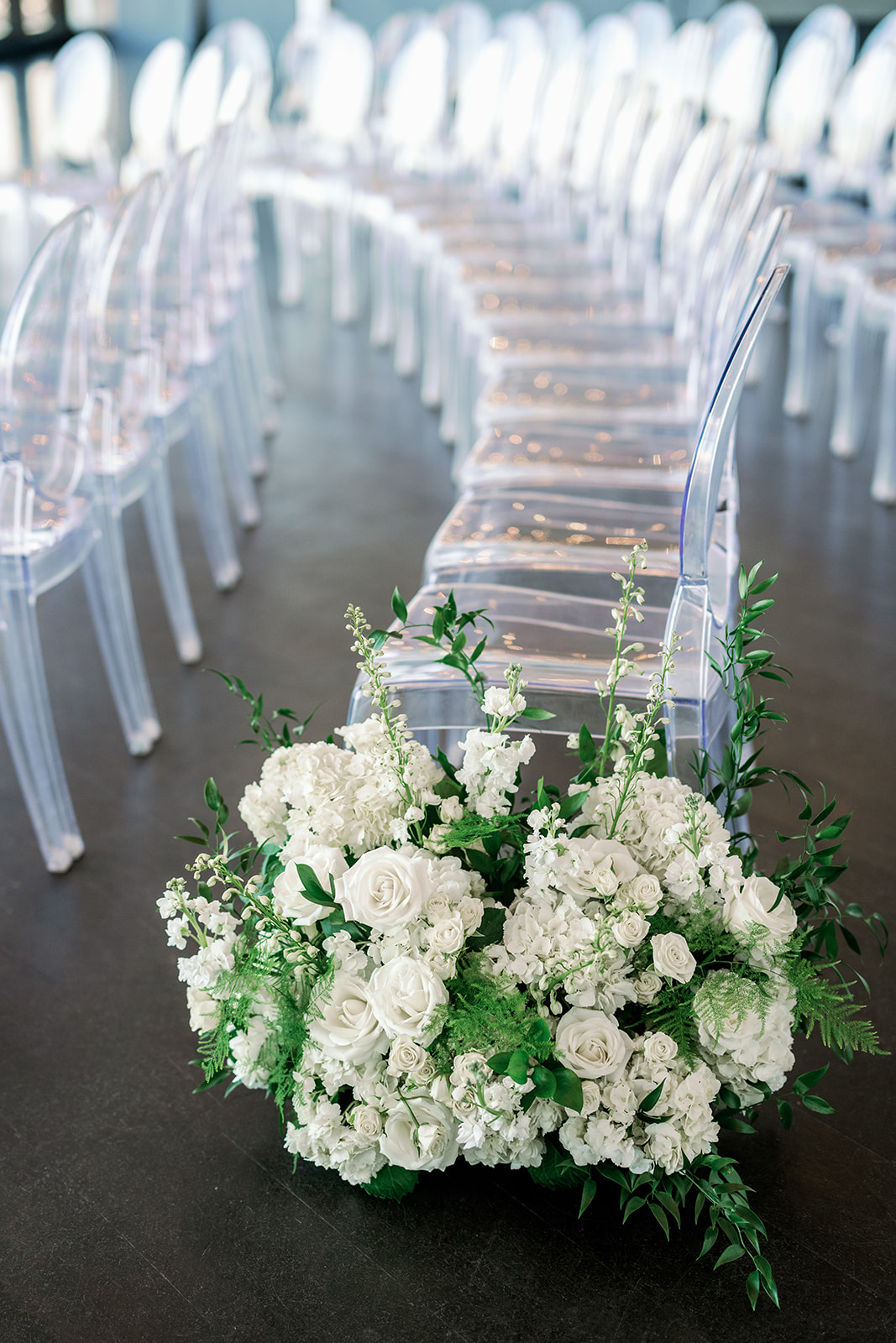 Indoor winter wedding ceremony aisle with white floral arrangements and ghost chairs at the Boston State Room.